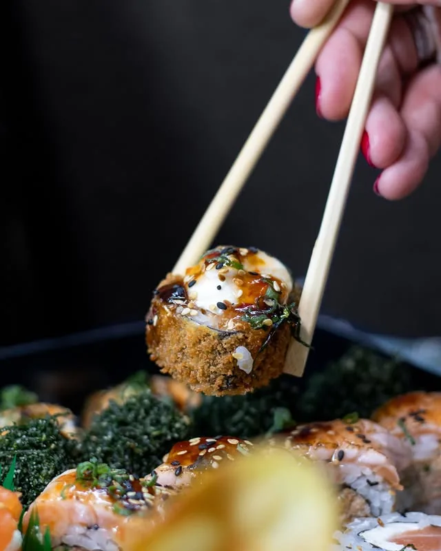 Peça de sushi empanado segurada por hashi sobre fundo escuro