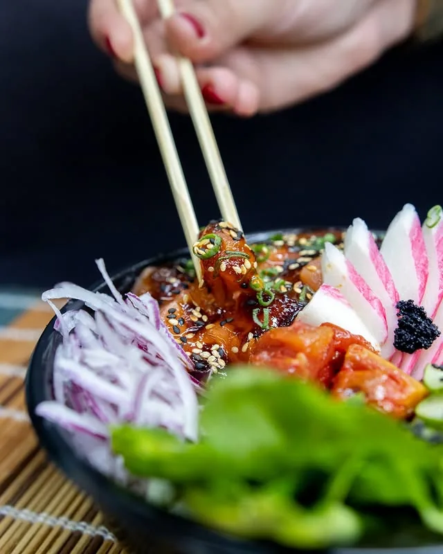 Tigela de poke com salmão, folhas e acompanhamentos sendo servida com hashi
