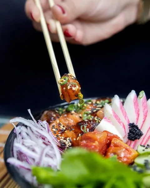 Tigela de poke com salmão sendo servida com hashi, com radish e folhas